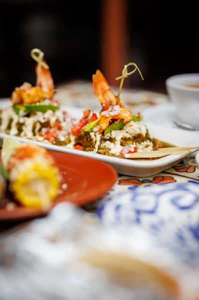 Elegant shrimp-topped enchiladas drizzled with crema and garnished with avocado, tomato, and cotija cheese, served on a rectangular plate over colorful tile.