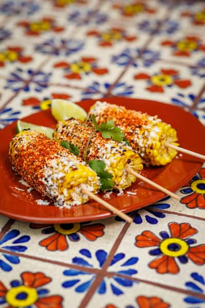 Authentic Mexican street corn (elotes) on wooden skewers, coated in cotija cheese, chili powder, and crema, served with lime wedges on a red plate atop a hand-painted Talavera tile table.