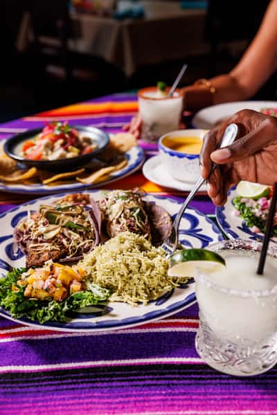 Colorful Mexican plate with purple corn tacos, cilantro-lime rice, and fresh mango salsa, as a fork reaches in for a bite. The table is decorated with a vibrant serape cloth and surrounded by ceviche, queso dip, and margaritas.