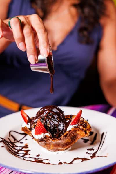 Close-up of a hand pouring rich chocolate sauce over a fried ice cream dessert, served in a crispy cinnamon shell with whipped cream and strawberries on a white plate, perfect for a sweet ending to a Mexican meal.