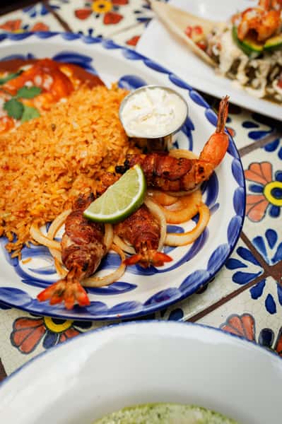 Grilled bacon-wrapped shrimp served on sautéed onions with a lime wedge, fluffy Mexican rice, and a side of crema on a blue-rimmed plate—perfect for a bold seafood entrée.
