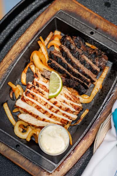 Sizzling fajita platter featuring grilled sliced chicken and charred steak strips over sautéed onions, served with a lime wedge and side of creamy sauce in a cast iron skillet on a wooden base.
