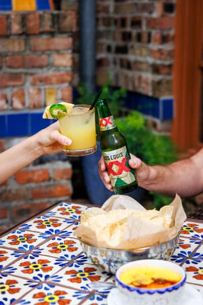 Cheers moment at a colorful Mexican restaurant, featuring a hand holding a pineapple-jalapeño margarita and another holding a cold Dos Equis beer, with a bowl of tortilla chips and queso dip on a Talavera-tiled table.