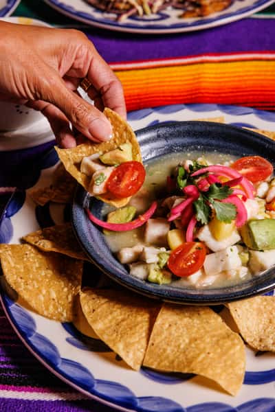 Hand scooping vibrant Mexican ceviche with tortilla chips, featuring chunks of white fish, avocado, cherry tomatoes, and pickled onions in a citrus marinade, served in a blue ceramic bowl.