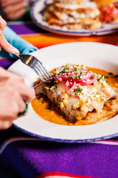 Close-up of hands cutting into cheesy enchiladas topped with pickled onions, cotija cheese, and cilantro, served over a rich red sauce.