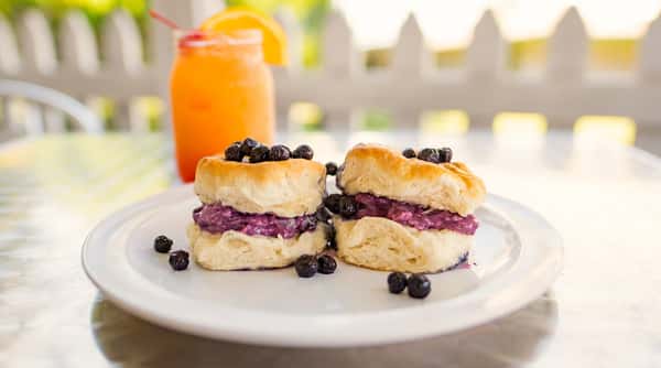 Two blueberry cream biscuits on a white plate with an orange juice in the background.