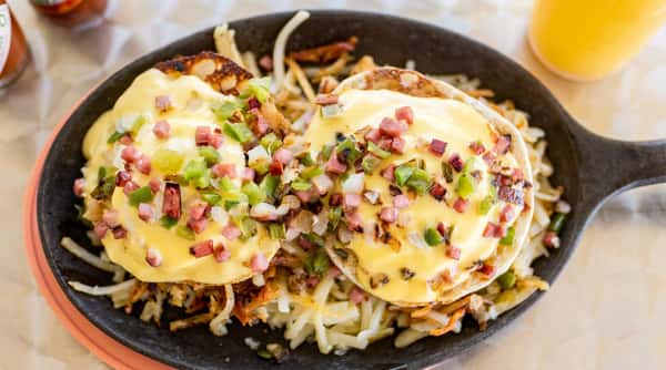 Eggs Benedict with hollandaise sauce, bell peppers, and ham on hash browns, served in a skillet.