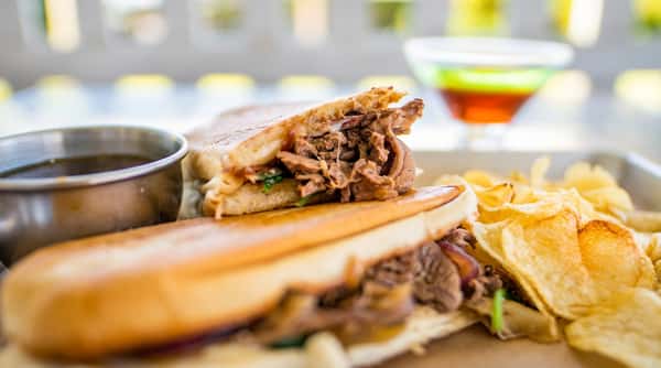 Pressed steak sandwich with caramelized onions, chips, and a dipping sauce on a metal tray.