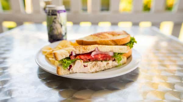 Chicken salad sandwich with lettuce, tomato, and onion on toasted bread, served with potato chips and a drink can on a metal table.