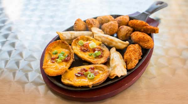 Appetizer platter on a cast iron pan with potato skins, dumplings, fried jalapeños, and hush puppies.