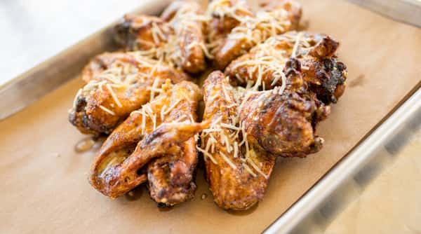 Baked chicken wings with brown crispy skin and shredded parmesan cheese on a brown paper-lined metal baking sheet.