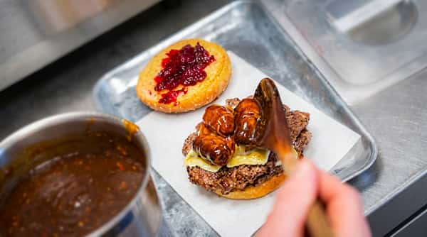 A burger being assembled: patty with cheese and gravy-covered tots, and a bun with red jam.