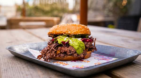 Pulled pork sandwich with green sauce and red cabbage on a sesame seed bun, served on a metal tray.