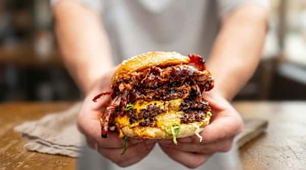 Hands holding a large double cheeseburger with pulled pork, bacon, and melted cheese on a sesame bun.