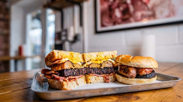 Breakfast sandwich with egg, bacon, black pudding, and a sausage roll with black pudding on a wooden board.