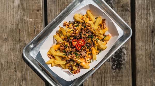 Fries topped with shredded meat, cheese sauce, red chili slices, and chives, served in a white paper tray on a metal serving tray.