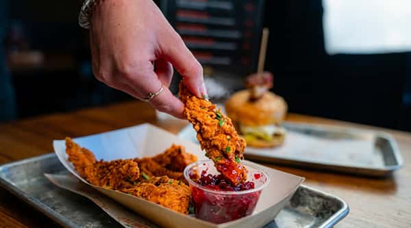 Hand dipping a crispy chicken tender into cranberry sauce, with more tenders and a burger in the background.