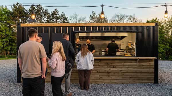 People queuing outside a black food truck with an open window showing chefs working inside.