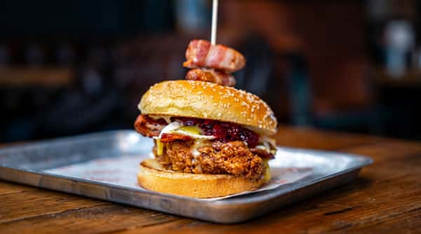 Fried chicken sandwich with bacon, cranberry sauce, and melted cheese on a toasted bun, garnished with bacon-wrapped onion rings.