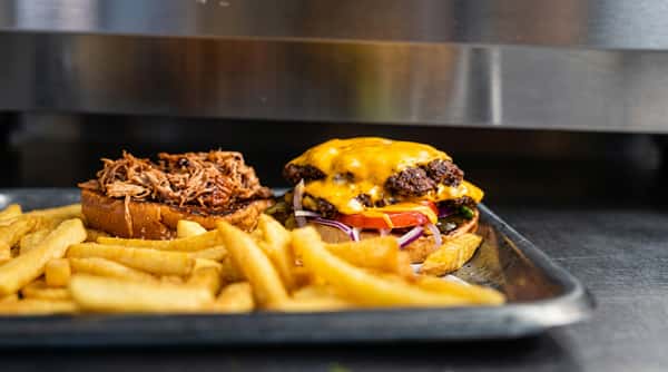 Cheeseburger with melted cheddar, tomato, onion, and fries, next to a pulled pork sandwich.