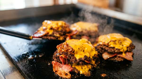 Four cheeseburger patties with melted yellow cheese and bacon cooking on a dark griddle.