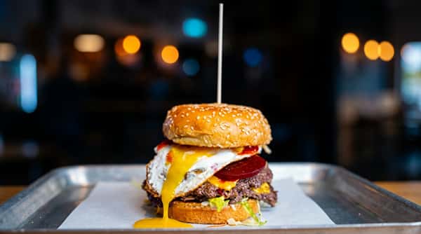 Beef burger with a fried egg, melted cheese, beets, and lettuce on a sesame seed bun, served on a tray.