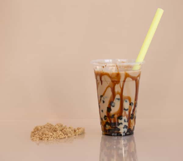 A cup of brown sugar boba tea with a yellow straw, sitting on a reflective surface with a pile of brown sugar beside it. This is one of the gallery images of Poke Tea.