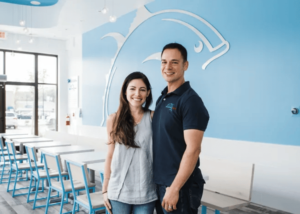 A light and airy restaurant setting, with a smiling couple standing in front of a blue wall featuring a large stylized fish logo. This restaurant is likely the subject of the "press" article being created in the site.
