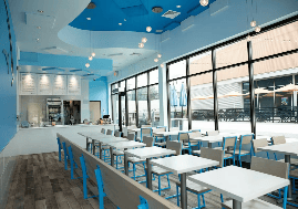 The interior of a restaurant with a blue color scheme has tables and chairs. Large windows let in natural light, and a row of menus is visible above a beverage cooler.