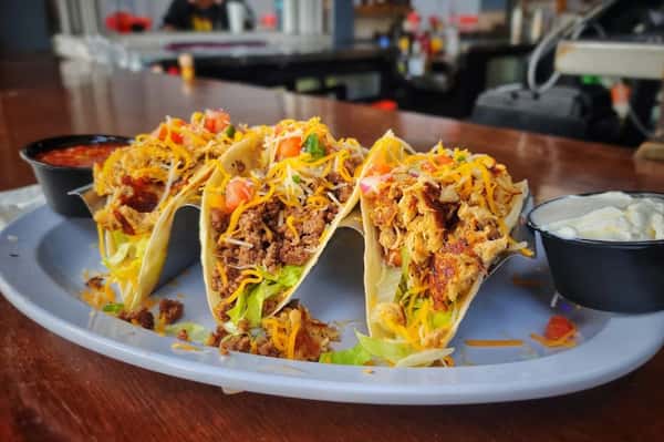 Three loaded tacos filled with seasoned beef and grilled chicken, topped with lettuce, cheese, and pico de gallo, served with salsa and sour cream.