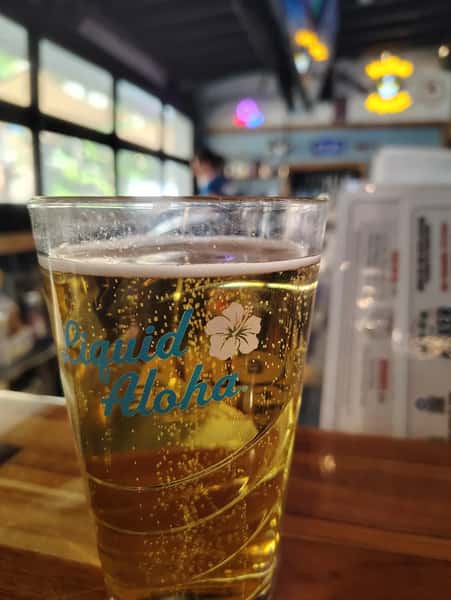 Golden draft beer served in a Liquid Aloha pint glass at a lively bar, featuring crisp carbonation and a relaxed coastal dining atmosphere.