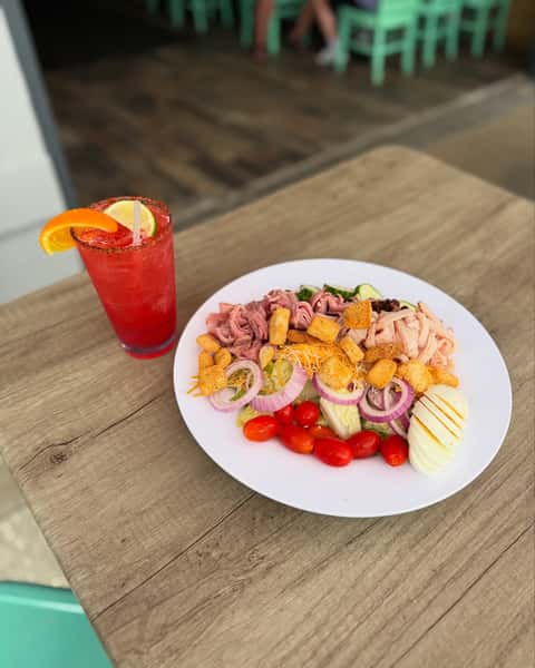 Chef salad topped with sliced ham, turkey, cheddar cheese, croutons, red onions, tomatoes, and hard-boiled egg, served with a vibrant red cocktail.