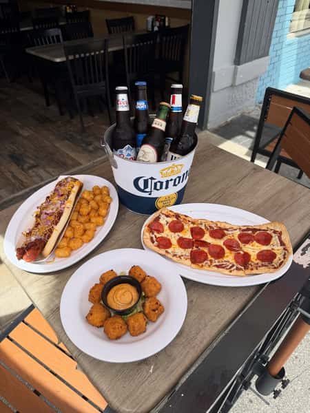 Assorted bar favorites including a loaded chili cheese hot dog with tater tots, pepperoni flatbread pizza, crispy seafood bites, and an ice-cold bucket of beer.
