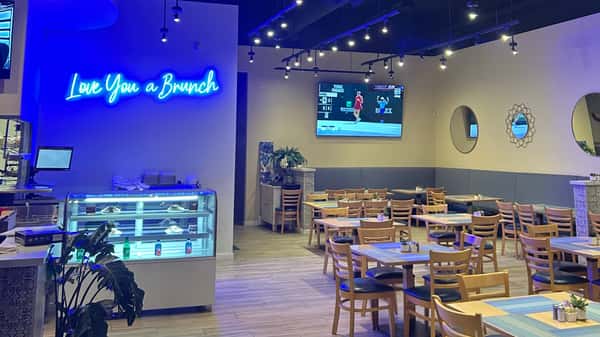 Interior dining room with wooden tables, chairs, and a blue neon sign reading "Love You a Brunch."