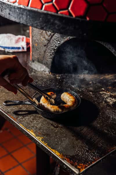 Close-up of Italian sausage sizzling in a cast iron skillet beside a rustic wood-fired oven, emphasizing traditional cooking methods and bold, smoky flavors in Italian cuisine.