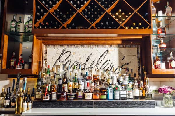 Stylish Italian restaurant bar with a large selection of spirits and wine bottles displayed on wood shelving and a custom mosaic "Punky's" sign above—perfect for showcasing a well‑stocked cocktail lounge atmosphere.