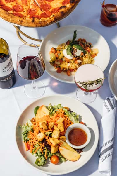 Close‑up of an Italian outdoor dining spread with red wine, a craft cocktail, pepperoni pizza, burrata pasta, and a crisp salad with apple and carrots on a white tablecloth in bright daylight, perfect for promoting alfresco dining and gourmet Italian fare.