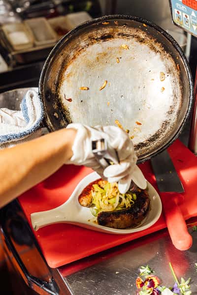 Grilled Italian sausage plated with caramelized onions in a rustic spoon dish, prepared in a professional kitchen setting—perfect for highlighting a hearty Italian entrée.