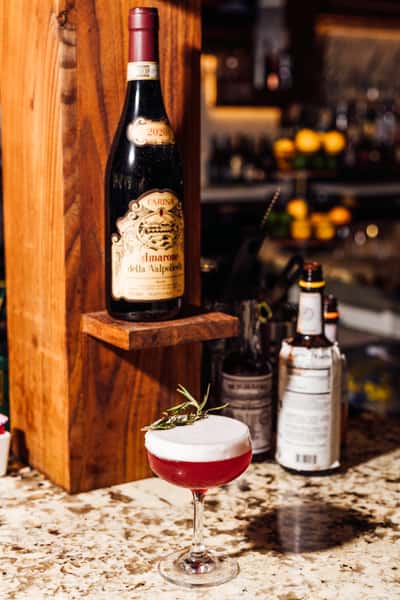 Vibrant red craft cocktail with a foamy top and rosemary garnish served on a granite bar, with a bottle of Amarone della Valpolicella wine displayed behind—perfect for promoting artisanal cocktails and Italian wine selections.
