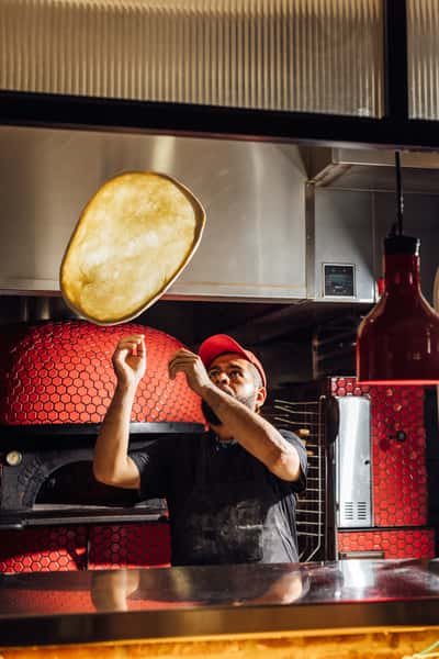 Chef tossing pizza dough high in the air in front of a traditional red-tiled wood-fired oven in an open kitchen setting.