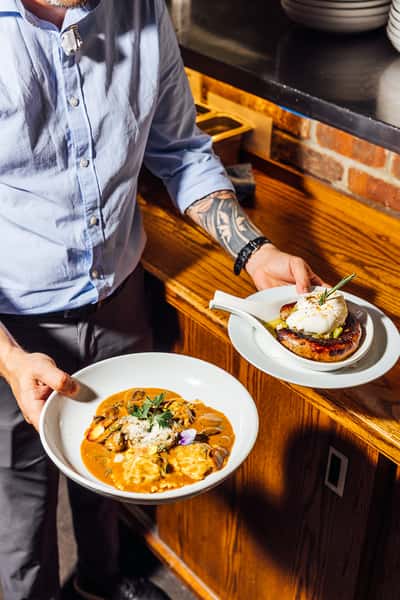 Waitstaff serving a gourmet Italian entrée of mushroom ravioli in a creamy tomato sauce topped with parmesan and herbs, alongside a grilled sausage appetizer finished with burrata and rosemary—perfect for showcasing upscale Italian dining.