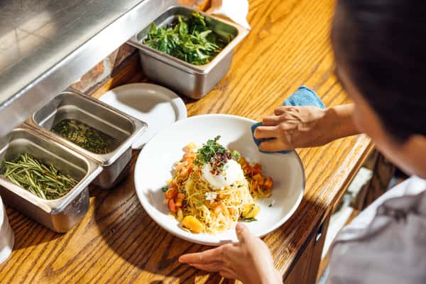 Freshly prepared pasta with diced tomatoes and creamy burrata cheese being plated and garnished with herbs at a kitchen pass.