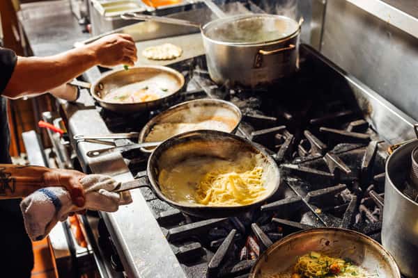 Italian chef cooking multiple pasta dishes over an open flame on a commercial kitchen stove, showcasing freshly made sauces and ingredients.