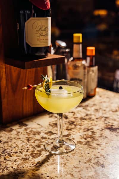 Elegant craft cocktail with a vibrant yellow hue served in a coupe glass, garnished with a rosemary sprig, lemon twist, and a single juniper berry, set on a granite bar counter with bottles of bitters and wine in the background—perfect for upscale bar or cocktail lounge ambiance.