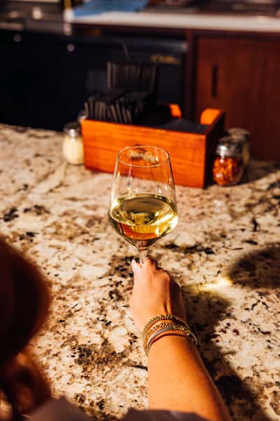 Close-up of a woman's hand holding a glass of chilled white wine at a granite bar counter, capturing the relaxed ambiance of a modern Italian restaurant or wine bar.
