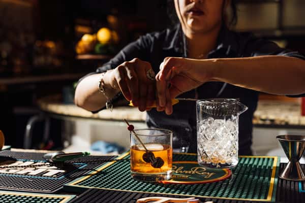 Bartender squeezing citrus peel into an old fashioned cocktail garnished with cherries at a well-lit bar counter.