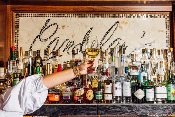 Woman raising a glass of white wine in front of a fully stocked bar featuring premium liquor bottles and a mosaic tile backdrop that reads "Punky's"—ideal for highlighting upscale bar offerings and vibrant cocktail lounge atmosphere.