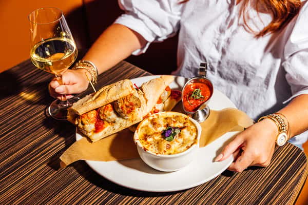 A woman seated at a table holding a glass of white wine, about to enjoy a meatball sub sandwich, baked cheesy pasta, and marinara sauce on a white plate.