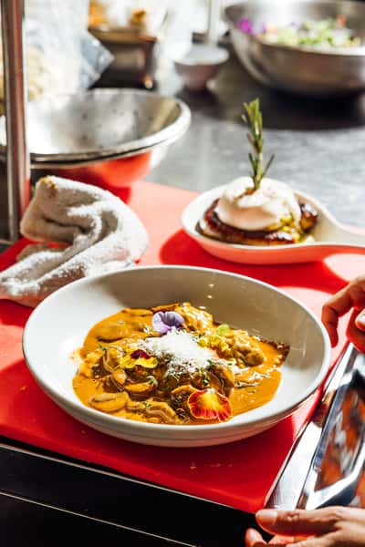 Close‑up of a plated creamy mushroom ravioli dish with shaved cheese and garnishes, alongside a burrata‑topped sausage in the background—ideal for showcasing gourmet Italian pasta dishes.