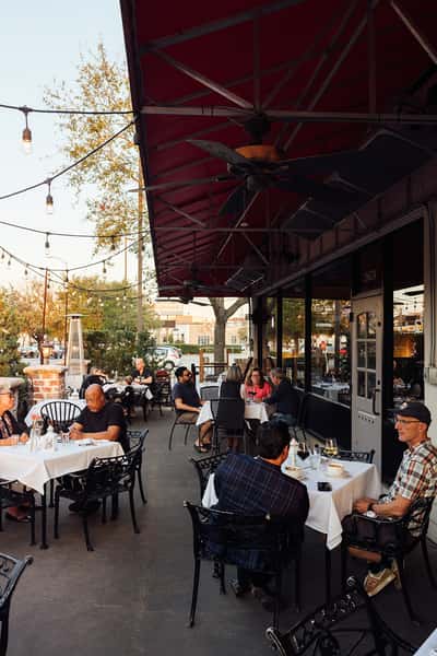 Diners enjoying meals on a charming outdoor patio with string lights, black wrought iron furniture, and a relaxed atmosphere at an Italian restaurant.